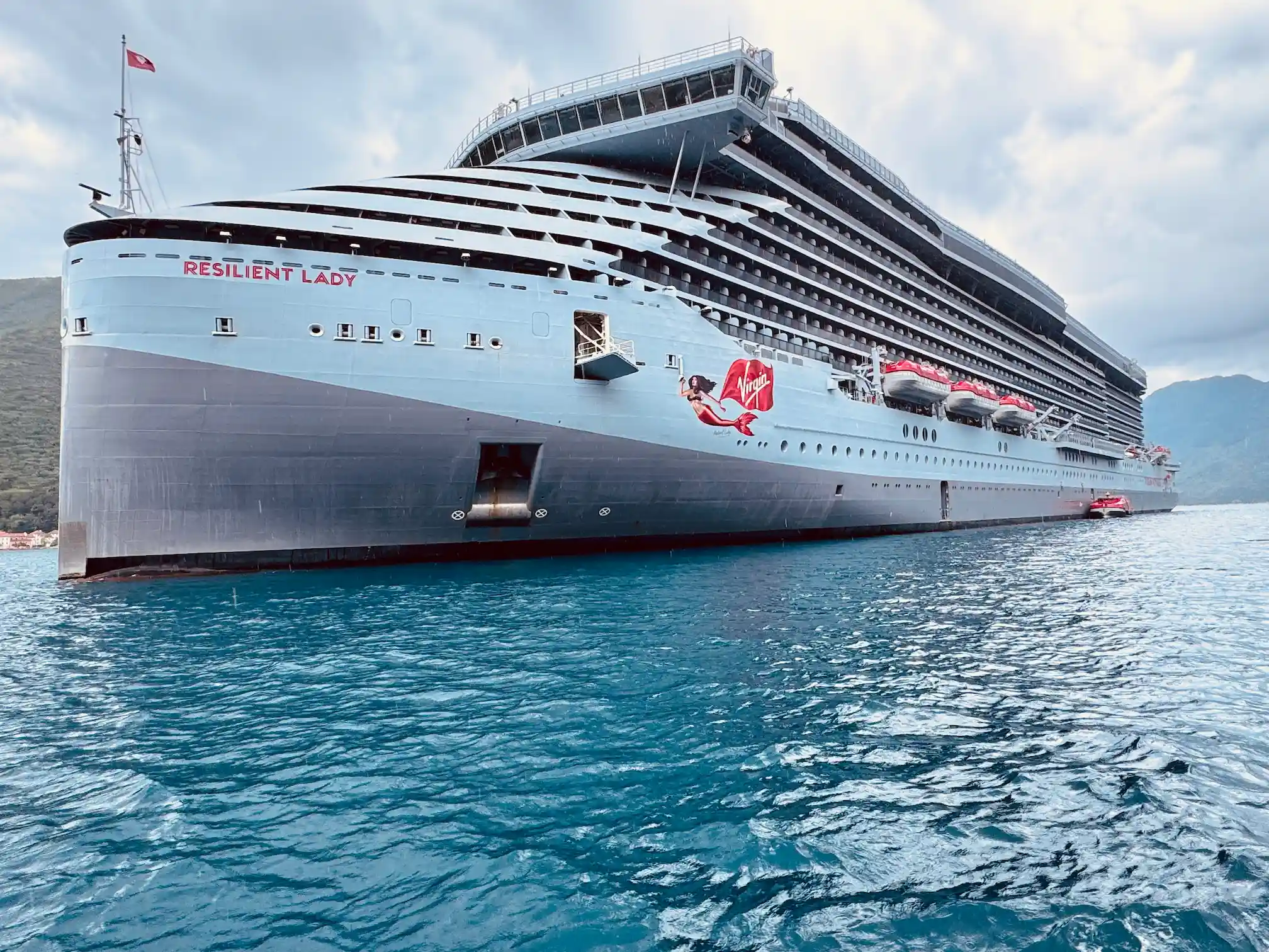 Virgin Voyages’ Resilient Lady cruise ship, mainly grey with bold red accents, sailing across calm clear waters beneath a cloudy sky.