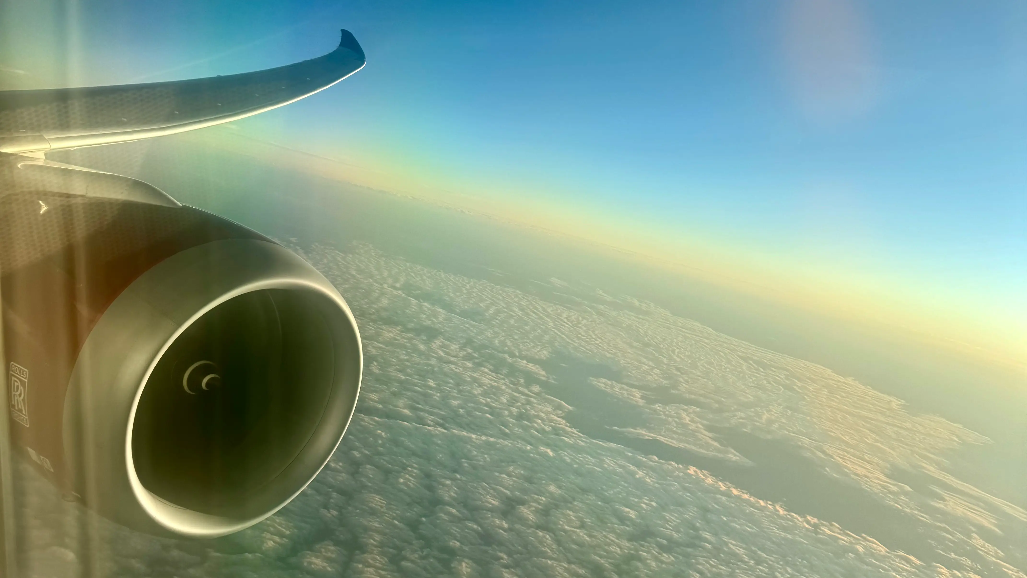 View from airplane window of a Rolls-Royce jet engine in flight, soaring above clouds with landscape visible below, capturing luxury air travel experience.